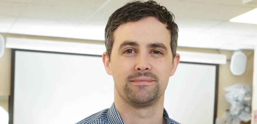 A close up of Gary McDonnell who is standing in front of a large pull down screen. He has short black hair and is wearing a dark blue checkered shirt.