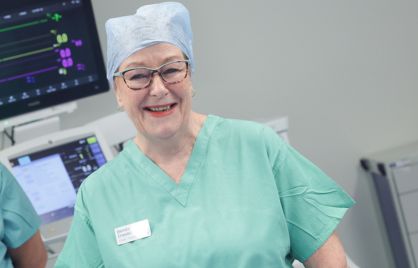 Deirdre Crosskill is smiling while standing in front of medical equipment in an operating theatre. She is wearing glasses, a green top and a blue theatre cap.