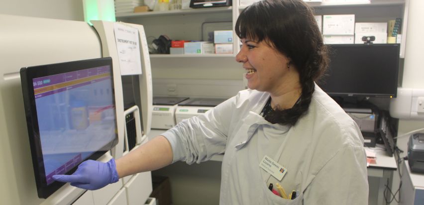 Maite is wearing a white lab coat and blue gloves as she presses a button on the touchscreen for one of the machines in the laboratory.