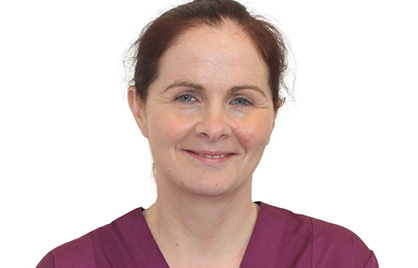 A headshot of Irene Murphy. She is smiling, has her hair tied back, and is standing against a white background and she is wearing a purple shirt.