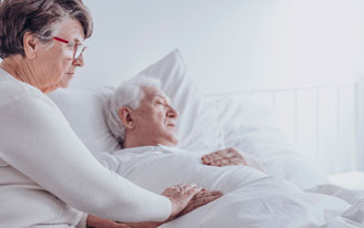 A woman sitting at the bedside of her husband who is asleep. She has her hand on his hand.