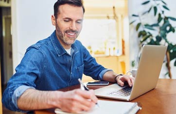 A man is sitting at a desk and is writing on a small notepad with one hand and using his laptop with the other.