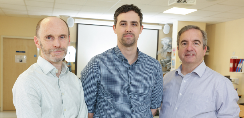 Three of the clinical engineers, Tom Bergin, Gary McDonnell and Glenn Tunney stand side by side in front of a large pulldown screen. Tom and Glen are wearing bright coloured shirts while Gary who is in the middle is wearing a dark blue checkered shirt.