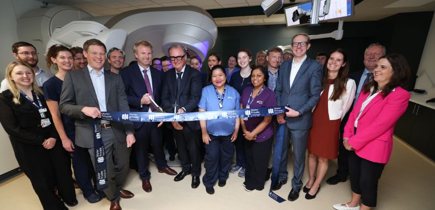 A group of people standing in front of a radiotherapy machine, cutting a ribbon