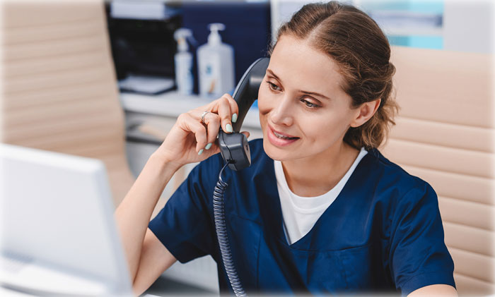 A woman is sitting in front of a computer screen and is holding a phone to her ear.