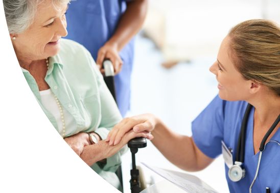 A female medical professional kneeling down and speaking with a female patient in a wheelchair.