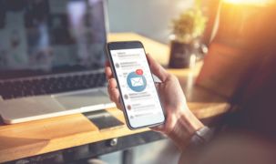 A person is sitting in front of a laptop and is holding up a phone which has an email icon on it.