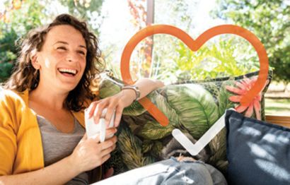 A woman is sitting on a chair holding a cup and is laughing. There is a heart animation beside her.
