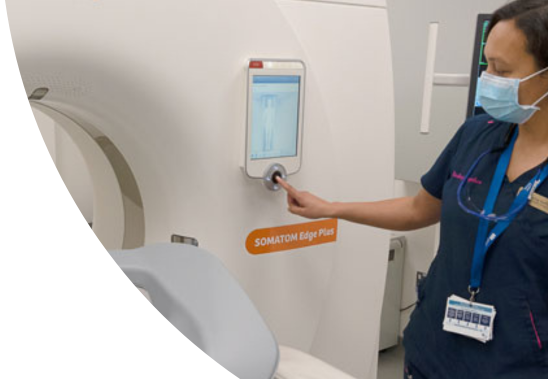 A woman who is wearing a blue uniform and a face mask is standing beside a CT machine and is pressing a button on the machine.